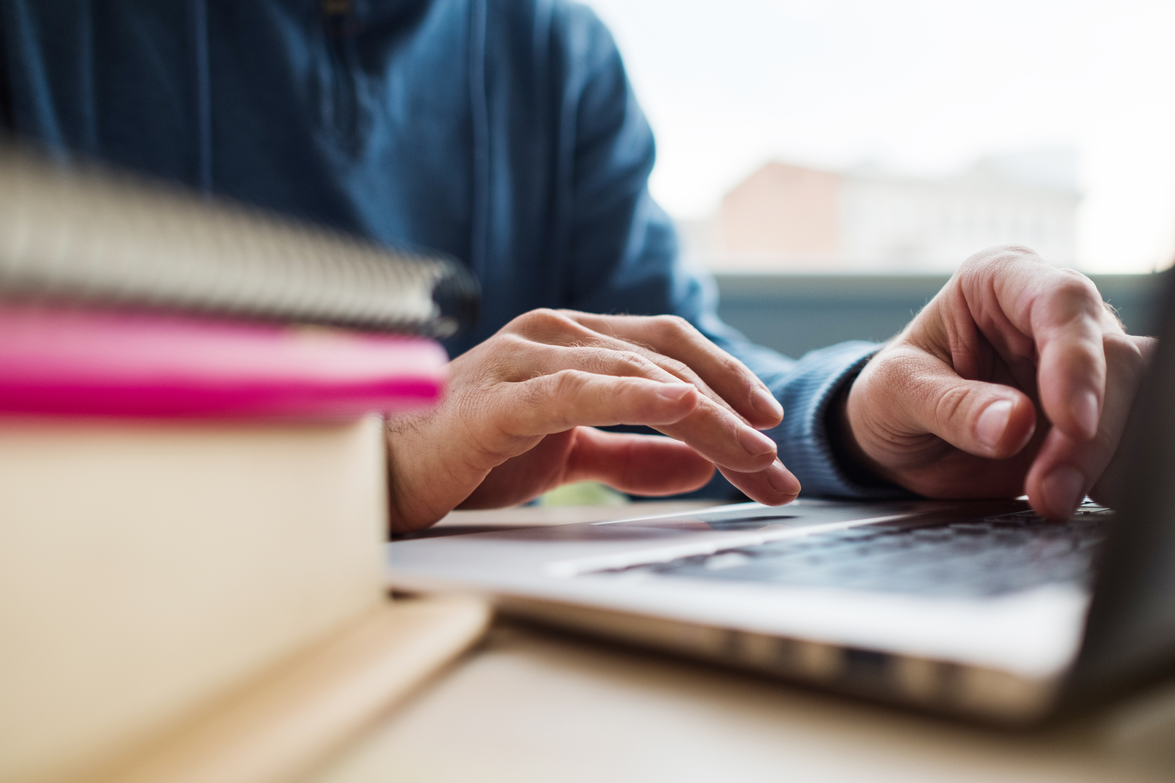 Close-up of hands typing on a laptop next to a stack of books and notebooks